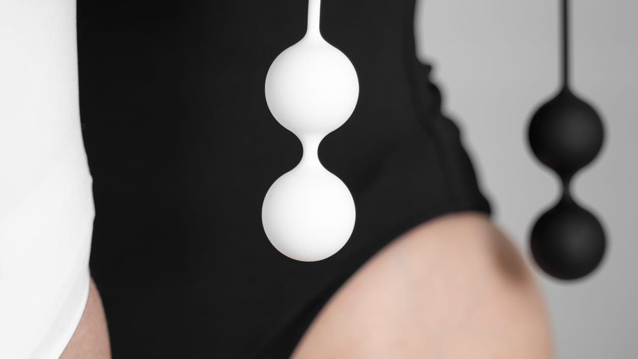 A person in a black bodysuit is shown, with a sharp close-up on their skin. Hanging prominently is a white double Ben Wa ball device, while a second black set is blurred in the background.