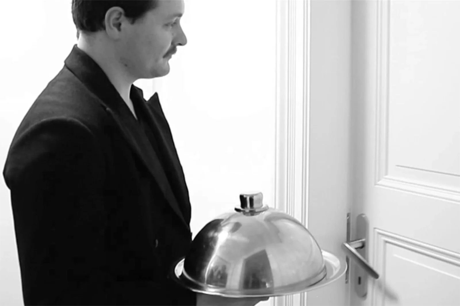 Black and white profile photo of a man in a dark suit holding a metal cloche tray by a door.