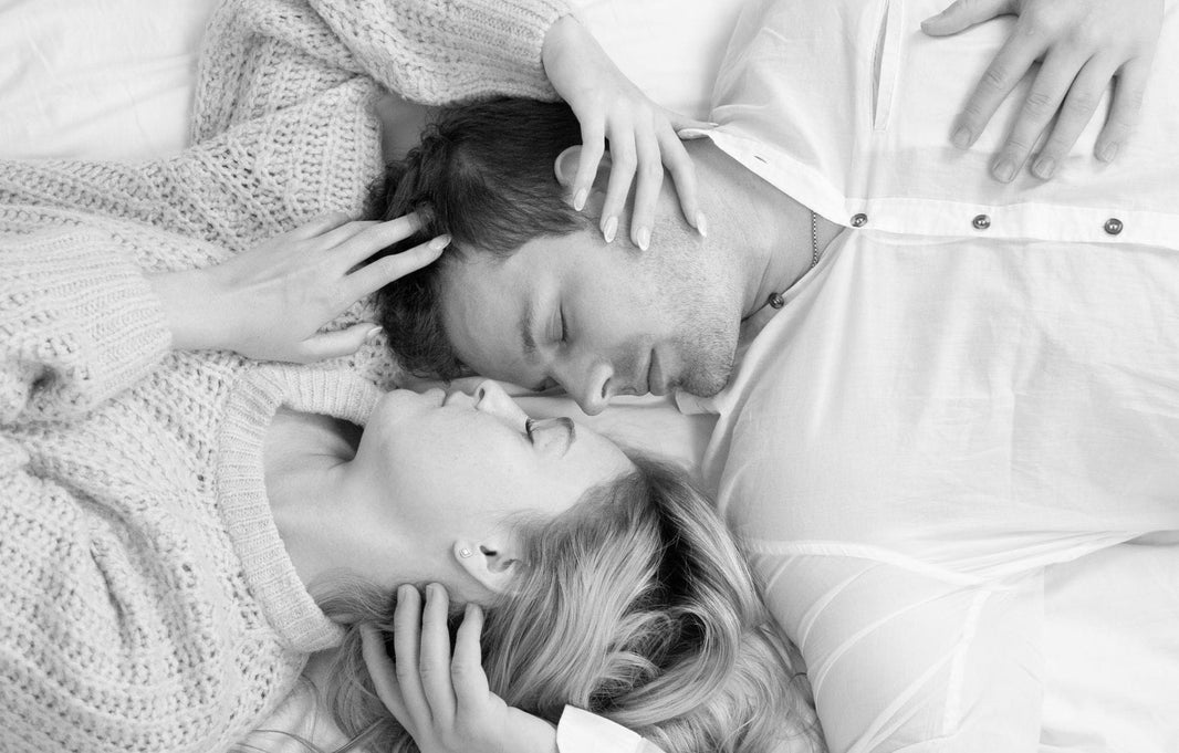 A black and white overhead photograph of a couple lying close together on a bed, faces nearly touching as they sleep, with hands gently caressing.