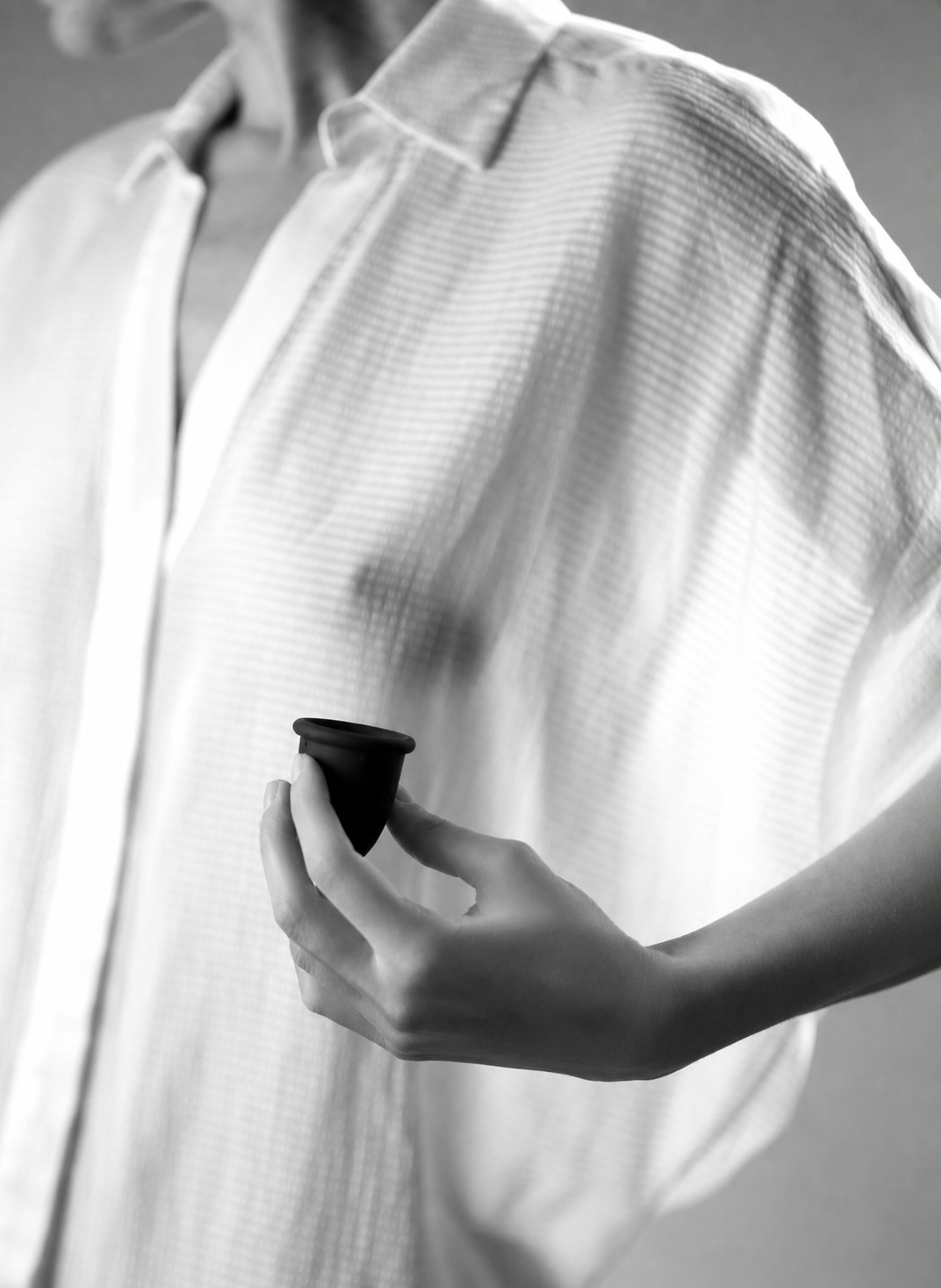 Artistic black and white mobile hero photo of a woman in a sheer white shirt holding a black Whoop-de-doo Noir menstrual cup.