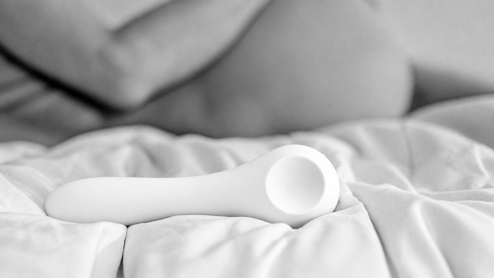 A luxury white Whoop-de-doo Classic vibrator resting on white crumpled bed sheets with a blurred silhouette of a woman in the background.