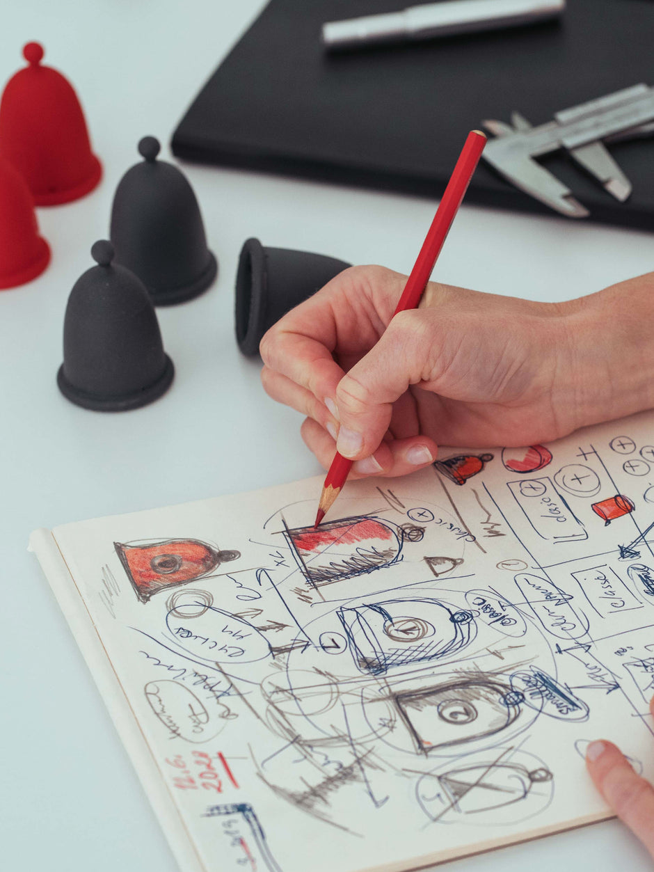 Close-up of a hand sketching anatomical details of menstrual cups in a notebook with a red pencil.