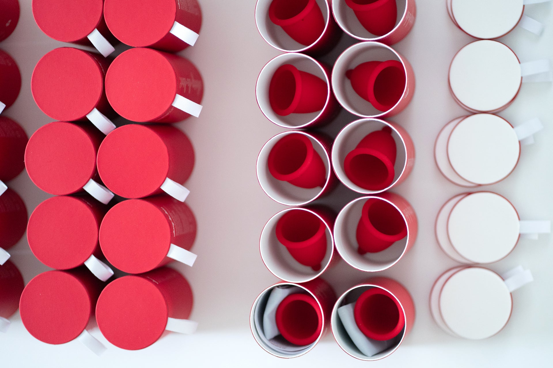 Symmetrical top-down view of red Whoop-de-doo menstrual cups inside protective paper tubes with pull-tab lids.