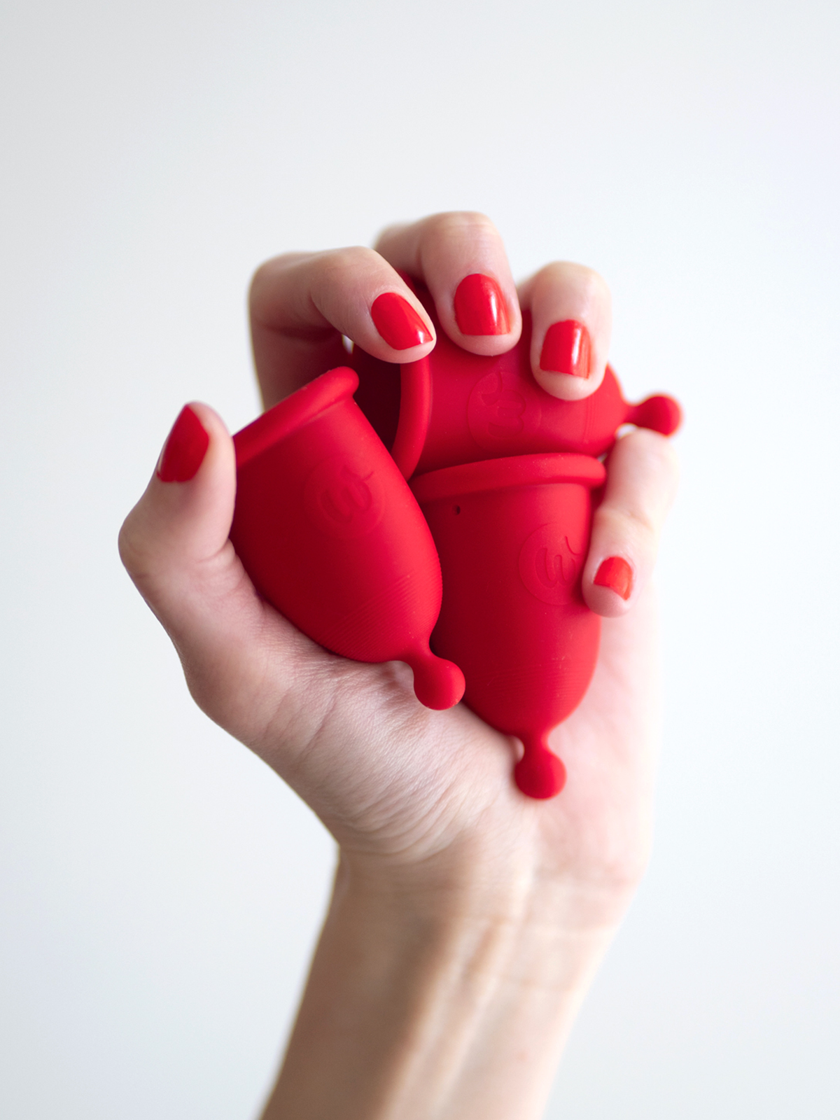 A woman’s hand with red nail polish squeezing flexible red Whoop-de-doo menstrual cups.