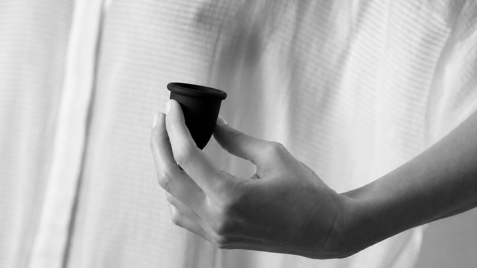 A minimalist close-up of a woman's hand showcasing the matte black Whoop-de-doo Noir menstrual cup against a soft, sheer white fabric.