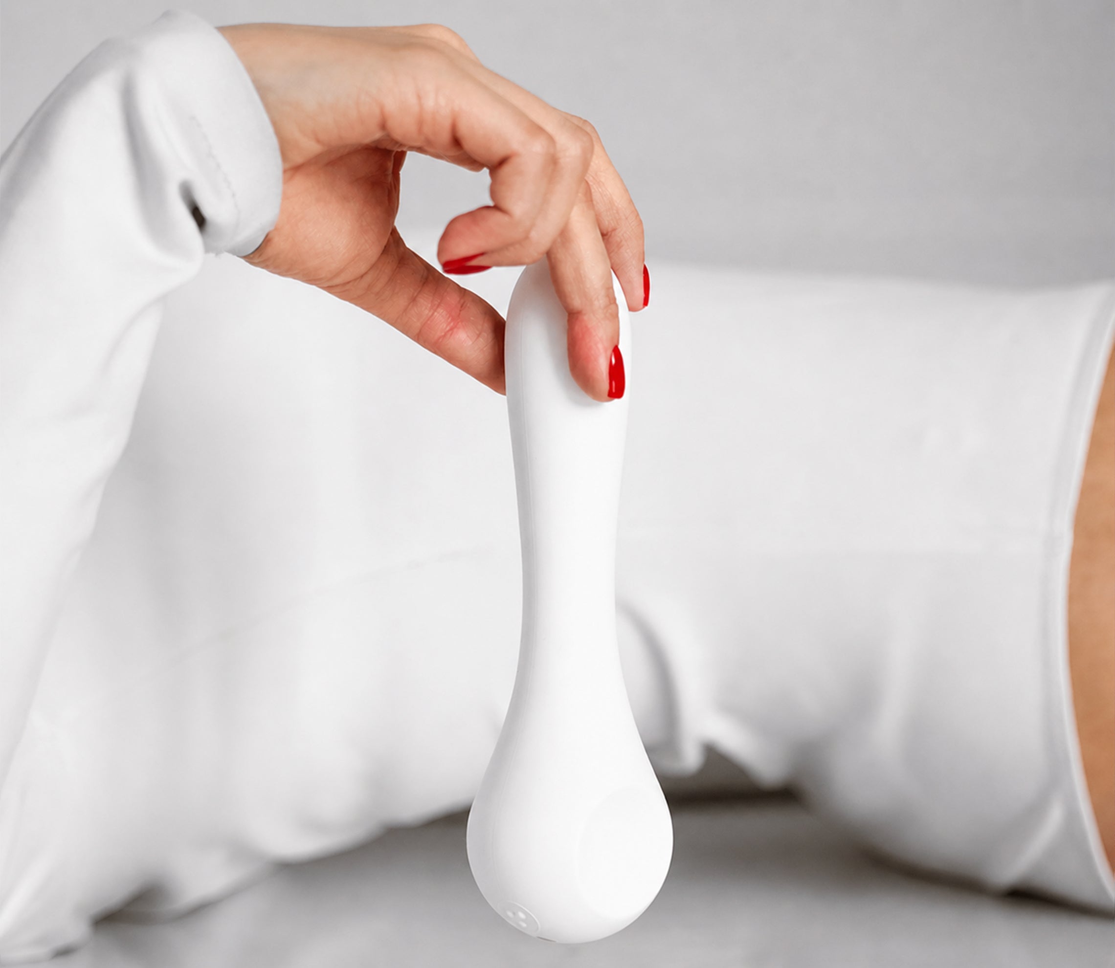 Close-up of a woman's hand with red nails holding the white design vibrator Whoop-de-doo Classic against a minimalist bright background.